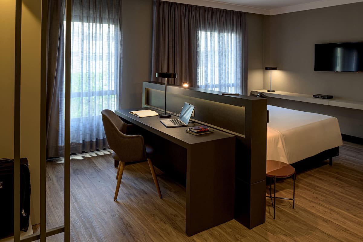 A hotel room with a desk, chair, bed, and a TV mounted on the wall.