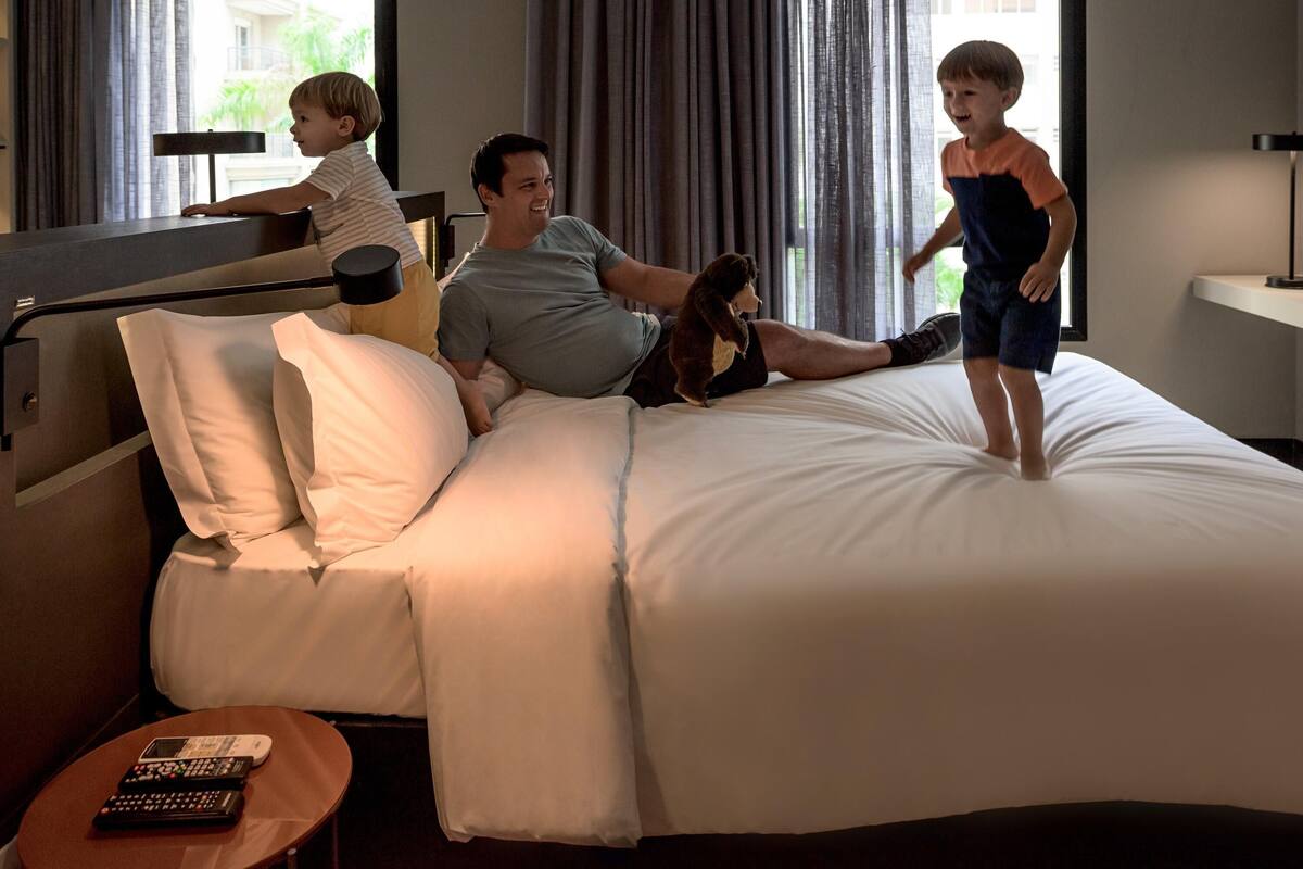 Ein Hotelzimmer mit Bett, Nachttisch mit Fernbedienung und Fenster mit Vorhängen.