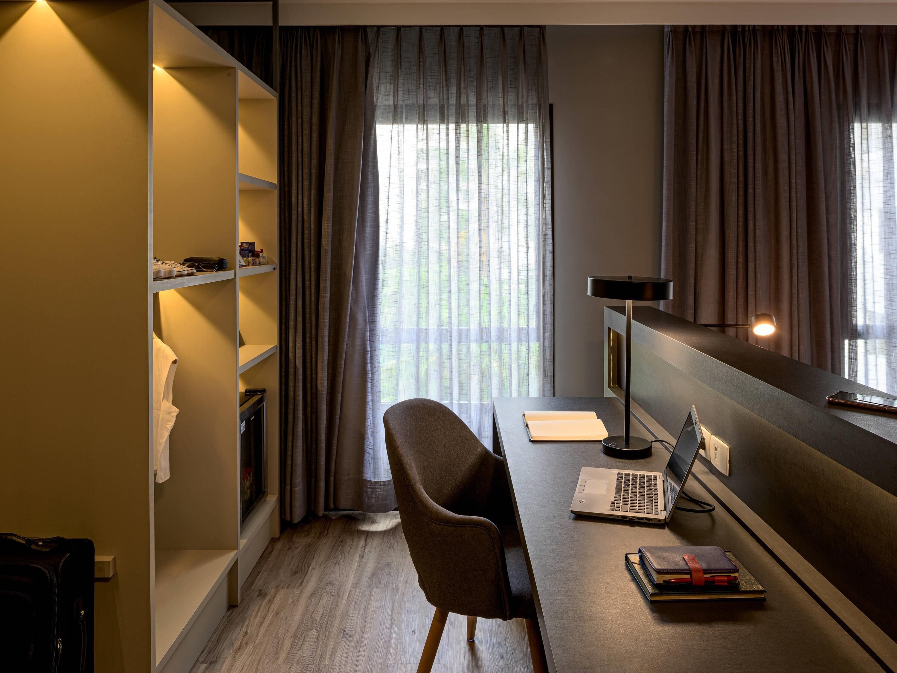 A modern room with a desk, laptop, and books. There is a window with curtains, a shelf with items, and a chair.