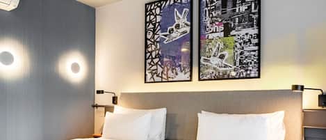 A hotel room with a double bed, two wall-mounted artworks, a ceiling air conditioner, and bedside lamps.