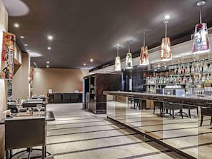 Uma área de bar com um balcão comprido, diversas luminárias pendentes e uma variedade de garrafas e copos nas prateleiras.