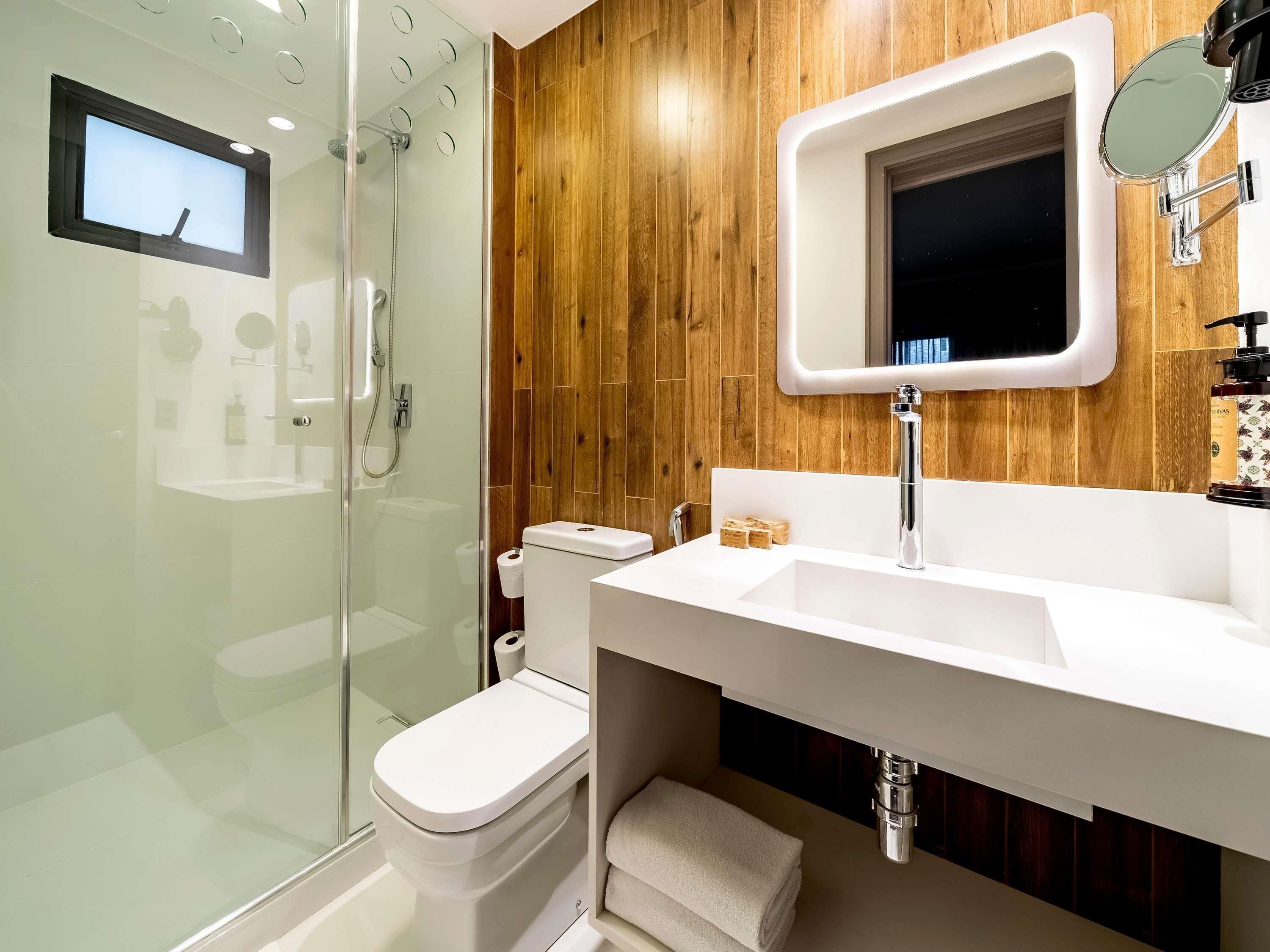 Une salle de bain moderne avec une douche à parois de verre, des toilettes blanches, un grand miroir et un lavabo blanc doté d’un distributeur de savon.