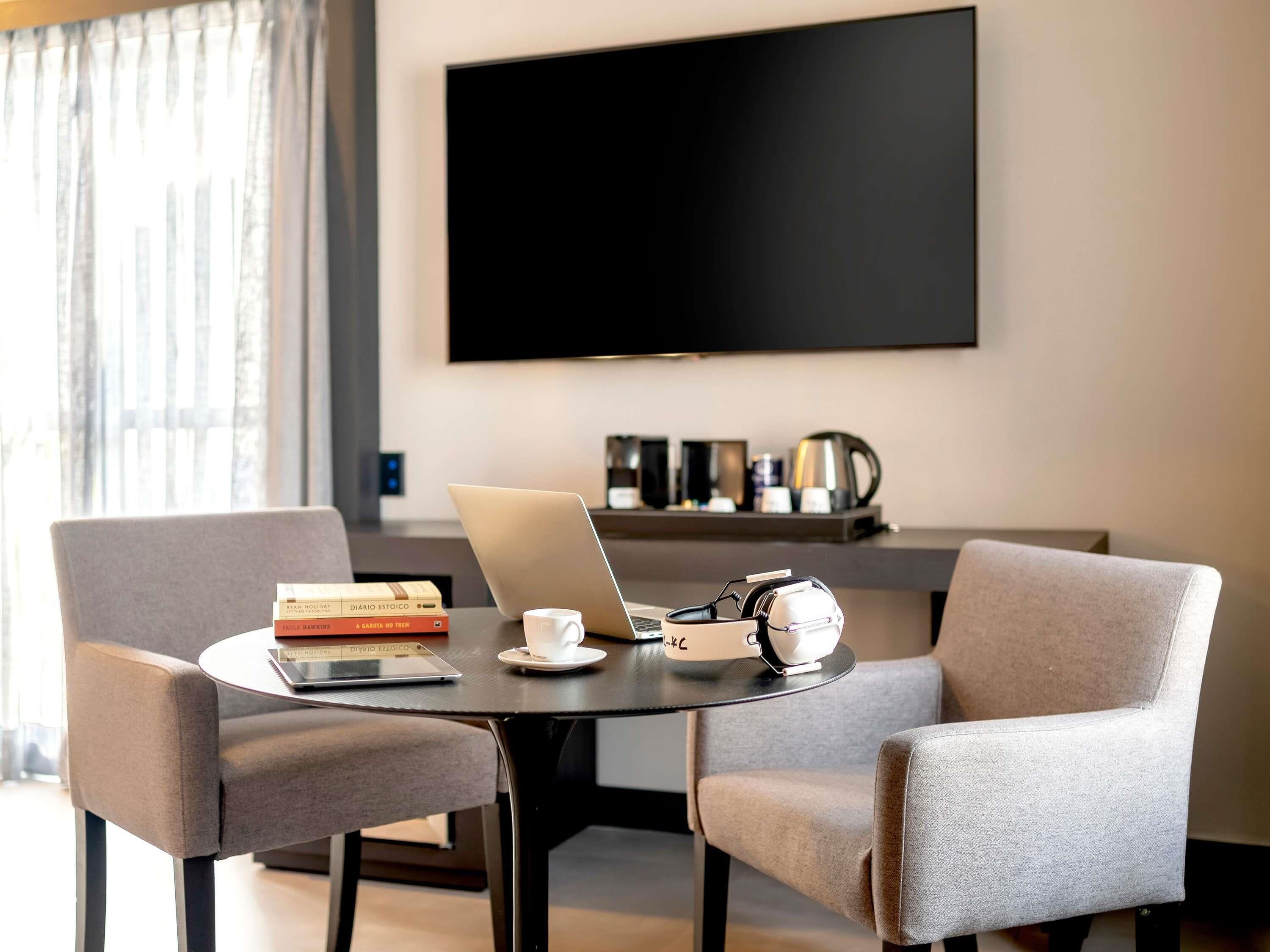 A modern hotel room with a round table, two grey armchairs, a laptop, a coffee machine, and a flat-screen TV mounted on the wall.