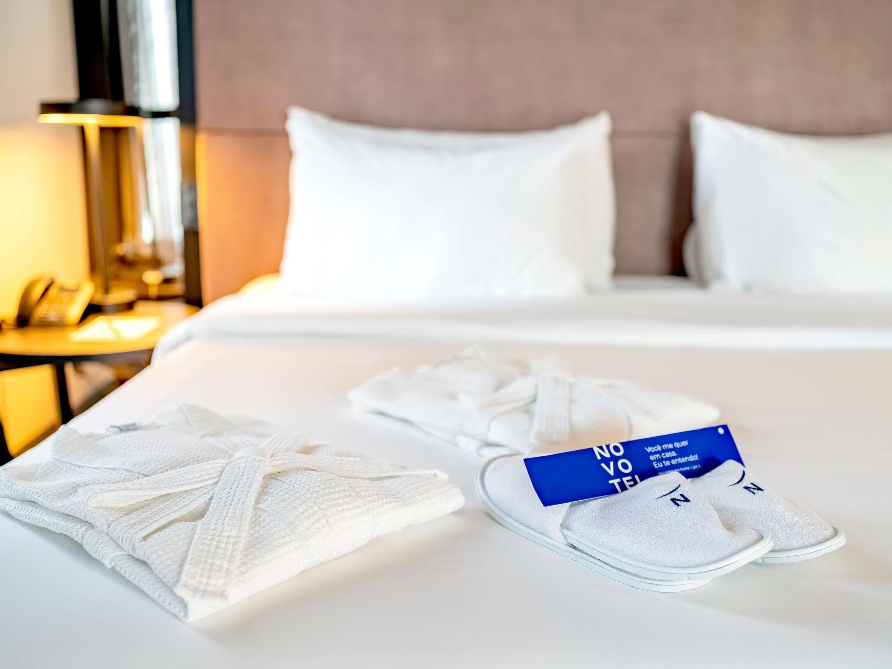 Een netjes opgemaakt bed met witte lakens, een paar witte sloffen en een blauwe kaart op het bed.