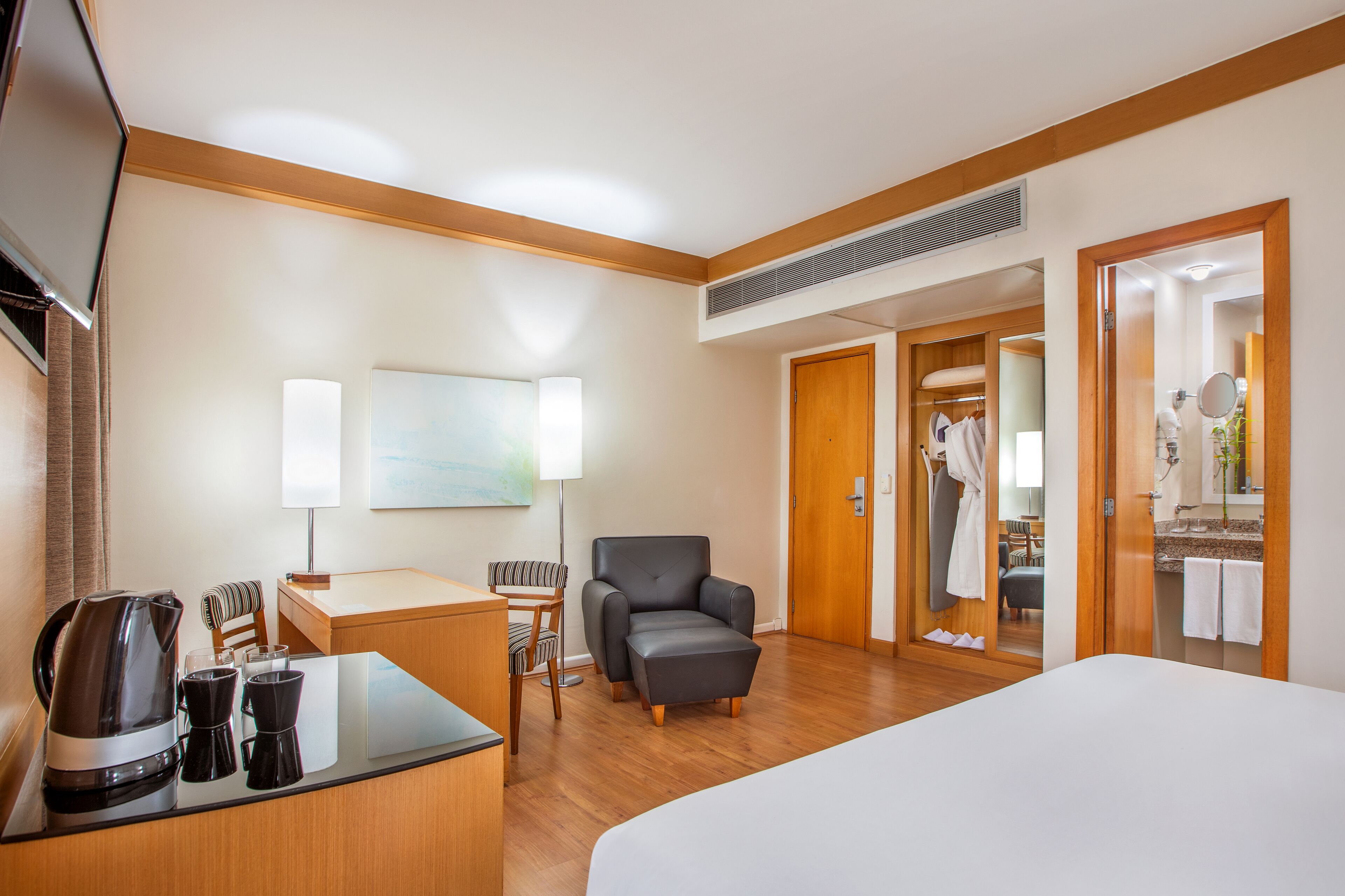 A hotel room with a bed, a desk with a coffee maker, a chair, a sofa, and a bathroom visible through an open door.