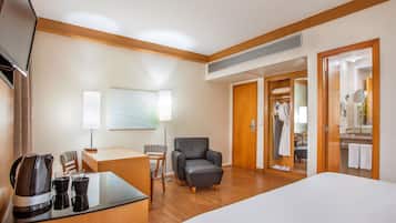 A hotel room with a bed, a desk with a coffee maker, a chair, a sofa, and a bathroom visible through an open door.