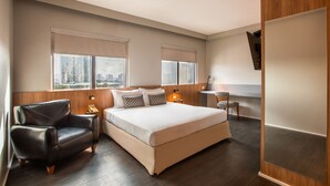 Una habitación de hotel moderna con una cama grande, dos mesitas de noche, un sillón de cuero y vista a la ciudad.