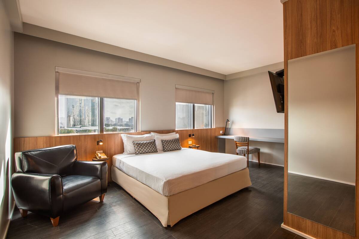 Una habitación de hotel moderna con una cama grande, dos mesitas de noche, un sillón de cuero y vista a la ciudad.