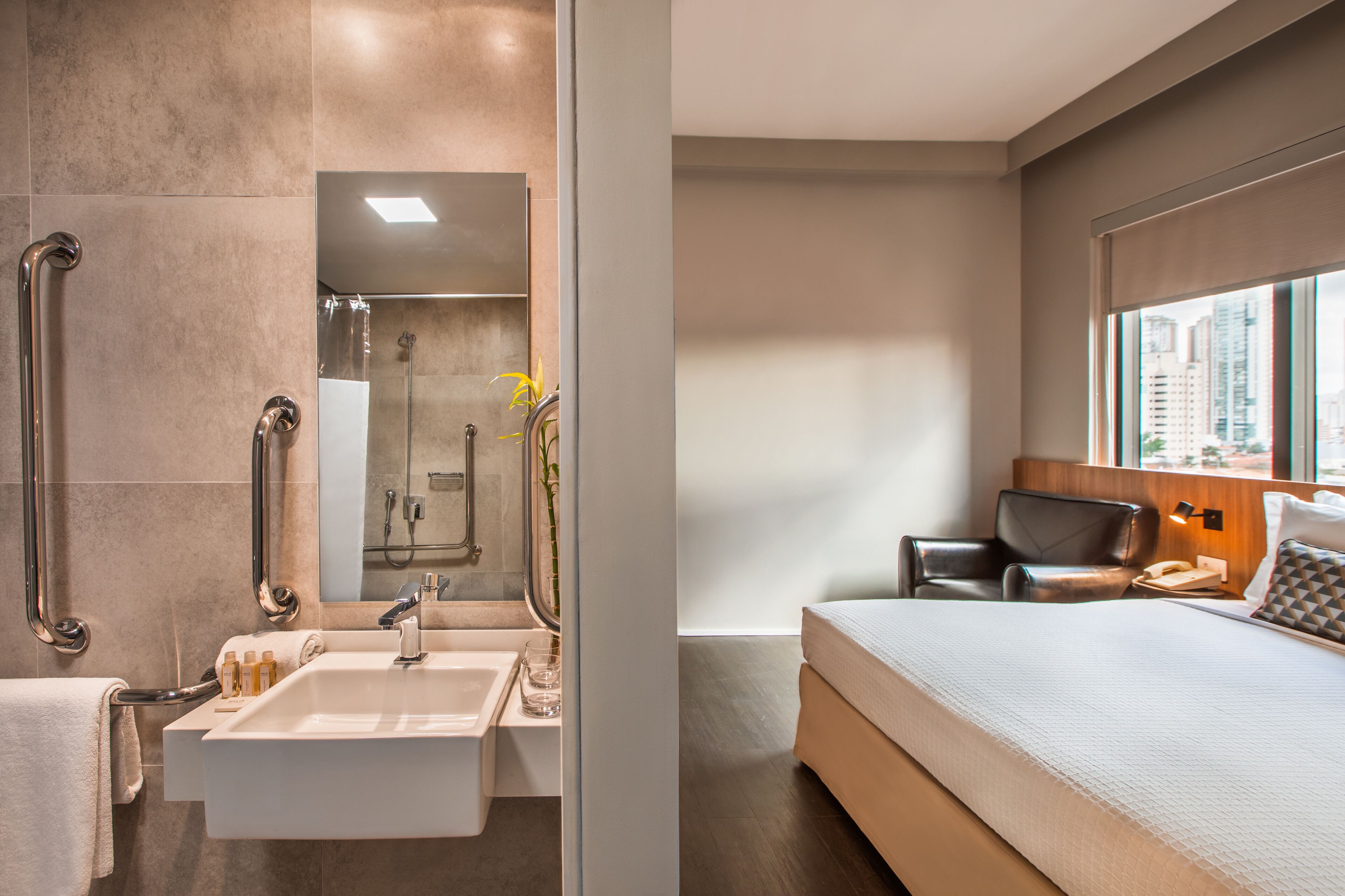 A modern bathroom with a sink, mirror, and shower, and a bedroom with a bed, nightstand, and window.