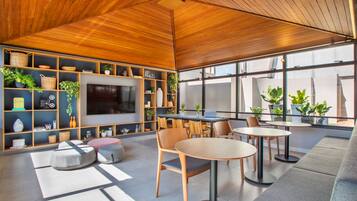 Un interior moderno con techo de madera, estanterías empotradas con plantas, un televisor de pantalla plana y zonas de estar con mesas y sillas.
