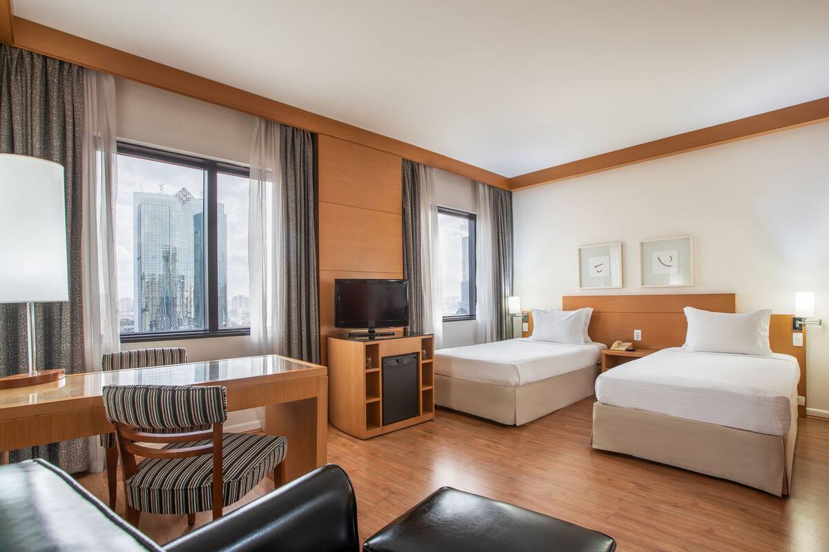 Una habitación de hotel con un escritorio de madera, dos camas, un televisor y ventanas amplias con cortinas.