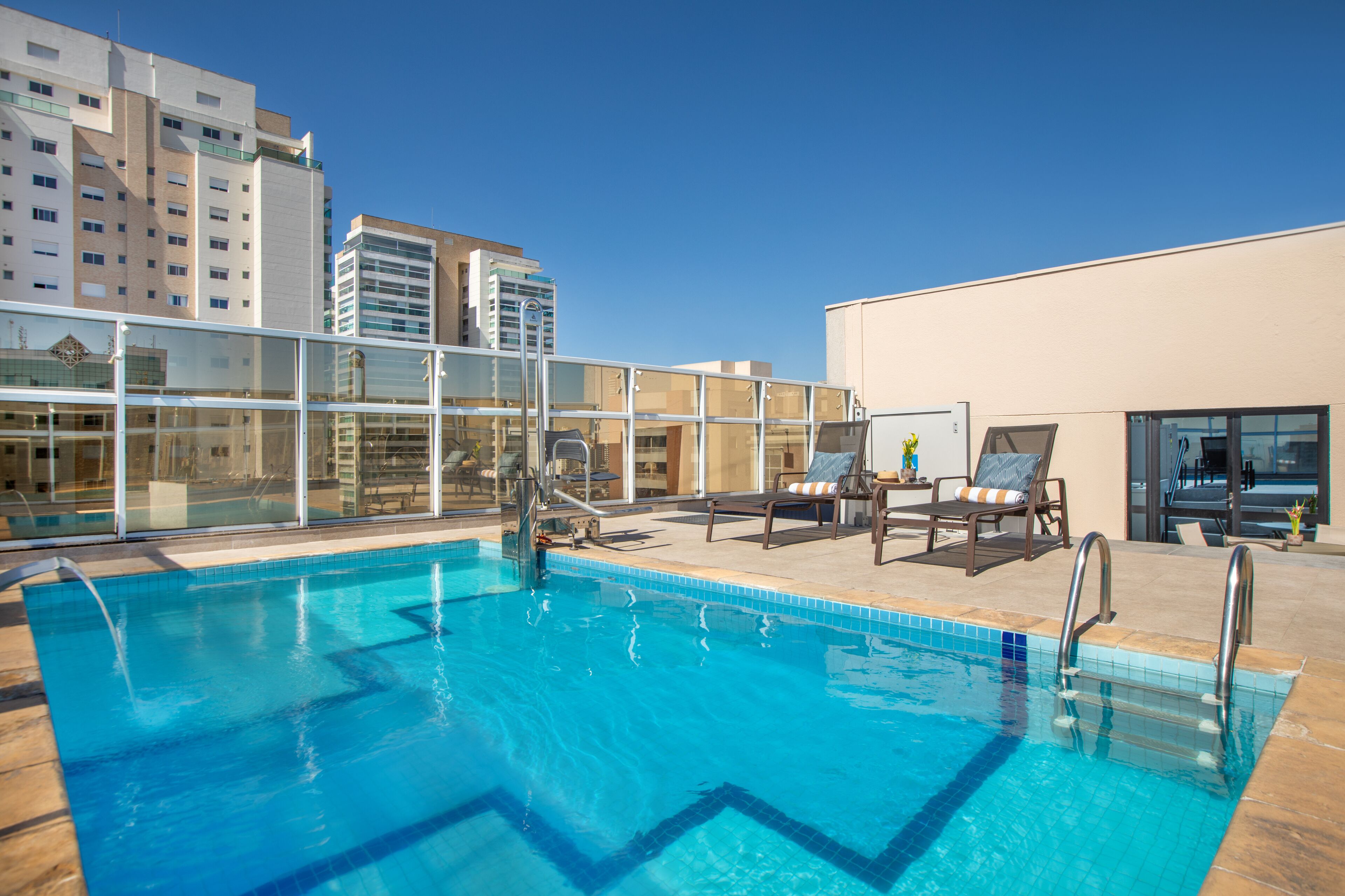 Piscina no terraço com água azul cristalina, rodeada por espreguiçadeiras e guarda-corpo de vidro.