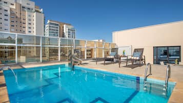 Piscina no terraço com água azul cristalina, rodeada por espreguiçadeiras e guarda-corpo de vidro.