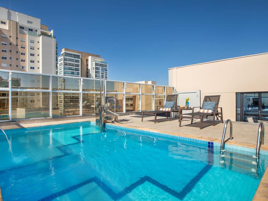 Piscina en la azotea con agua cristalina y azul, rodeada de sillones de descanso y una barandilla de vidrio.