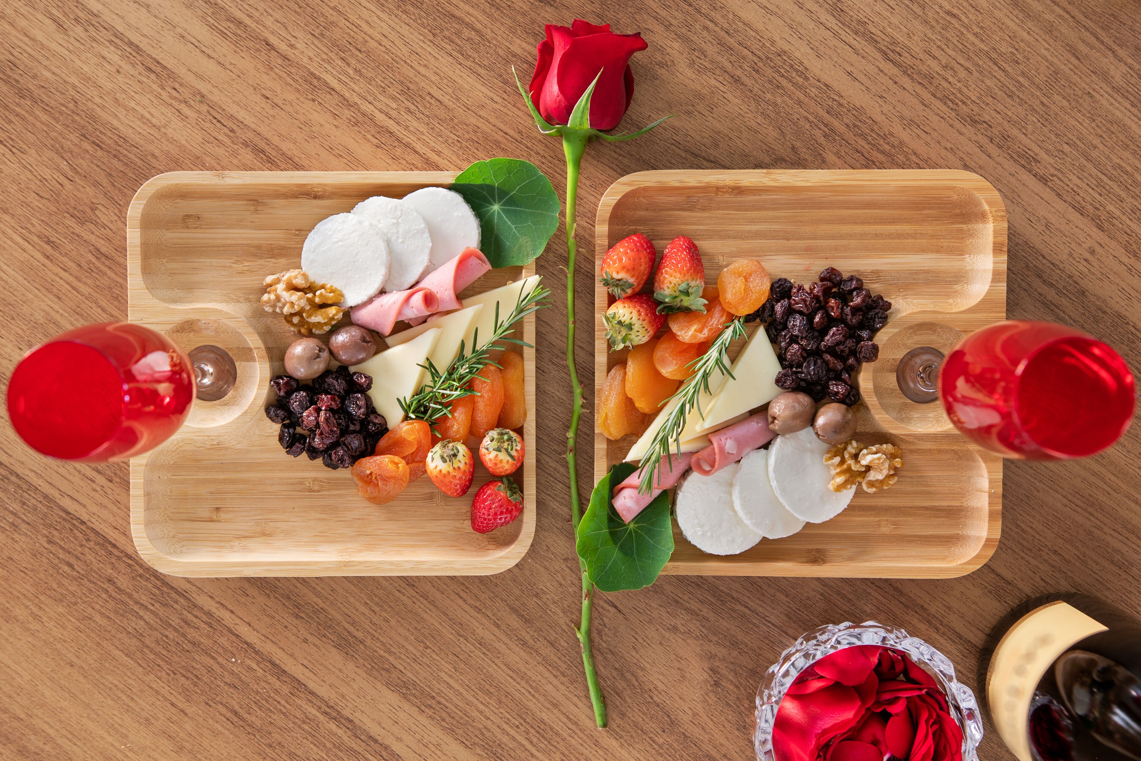 Two wooden serving boards with various cheeses, fruits, and nuts, accompanied by red wine glasses, on a wooden table.