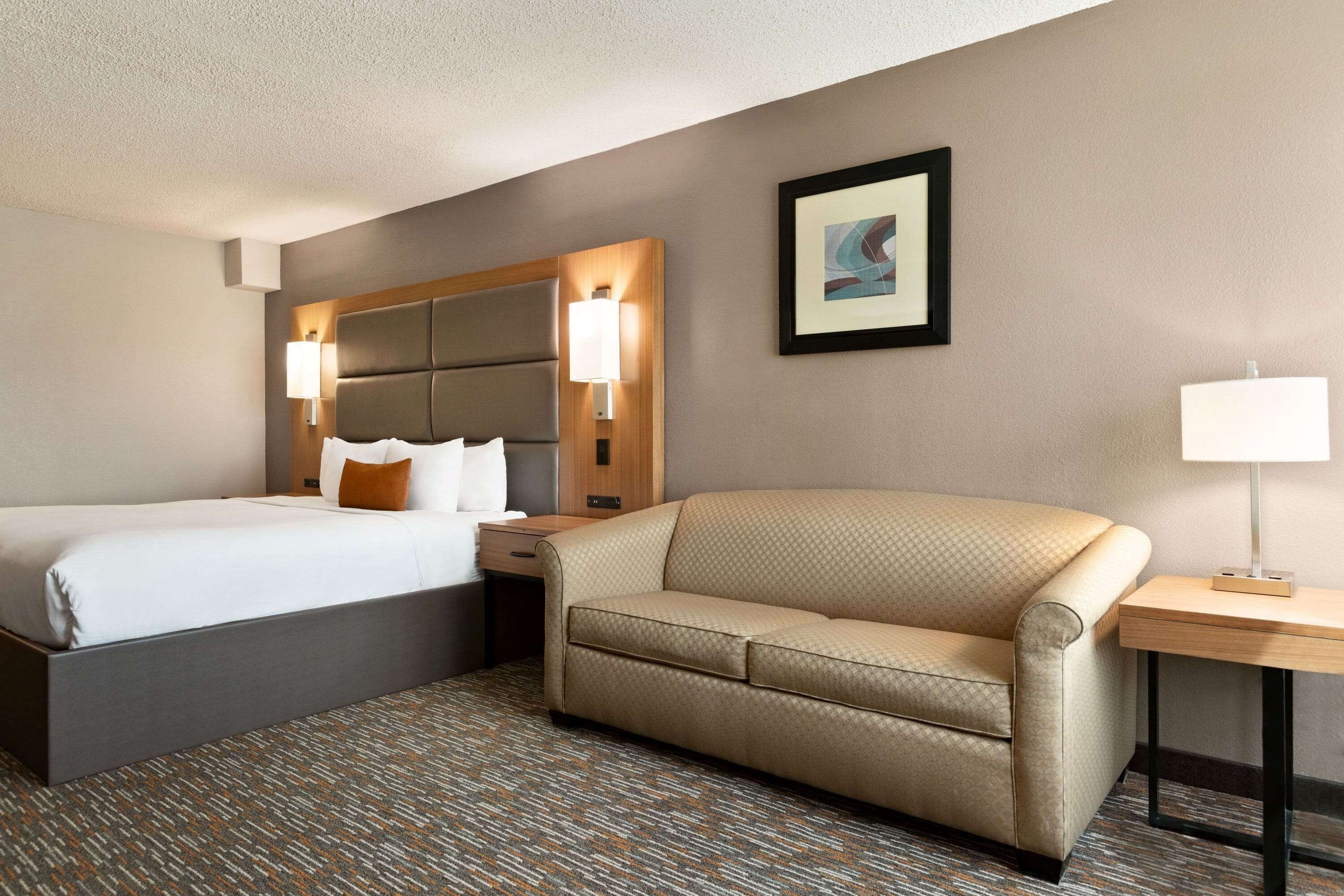 A hotel room with a bed, a sofa, a nightstand, a lamp, and a framed picture on the wall.