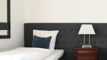 Een bed met witte beddengoed en een blauw kussen, een donker hoofdeinde, een nachtkastje met een lamp, en een houten nachtkastje.