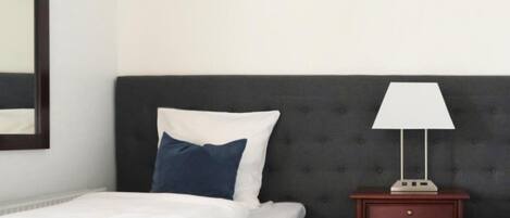 A bed with white bedding and a blue pillow, a dark headboard, a bedside table with a lamp, and a wooden nightstand.