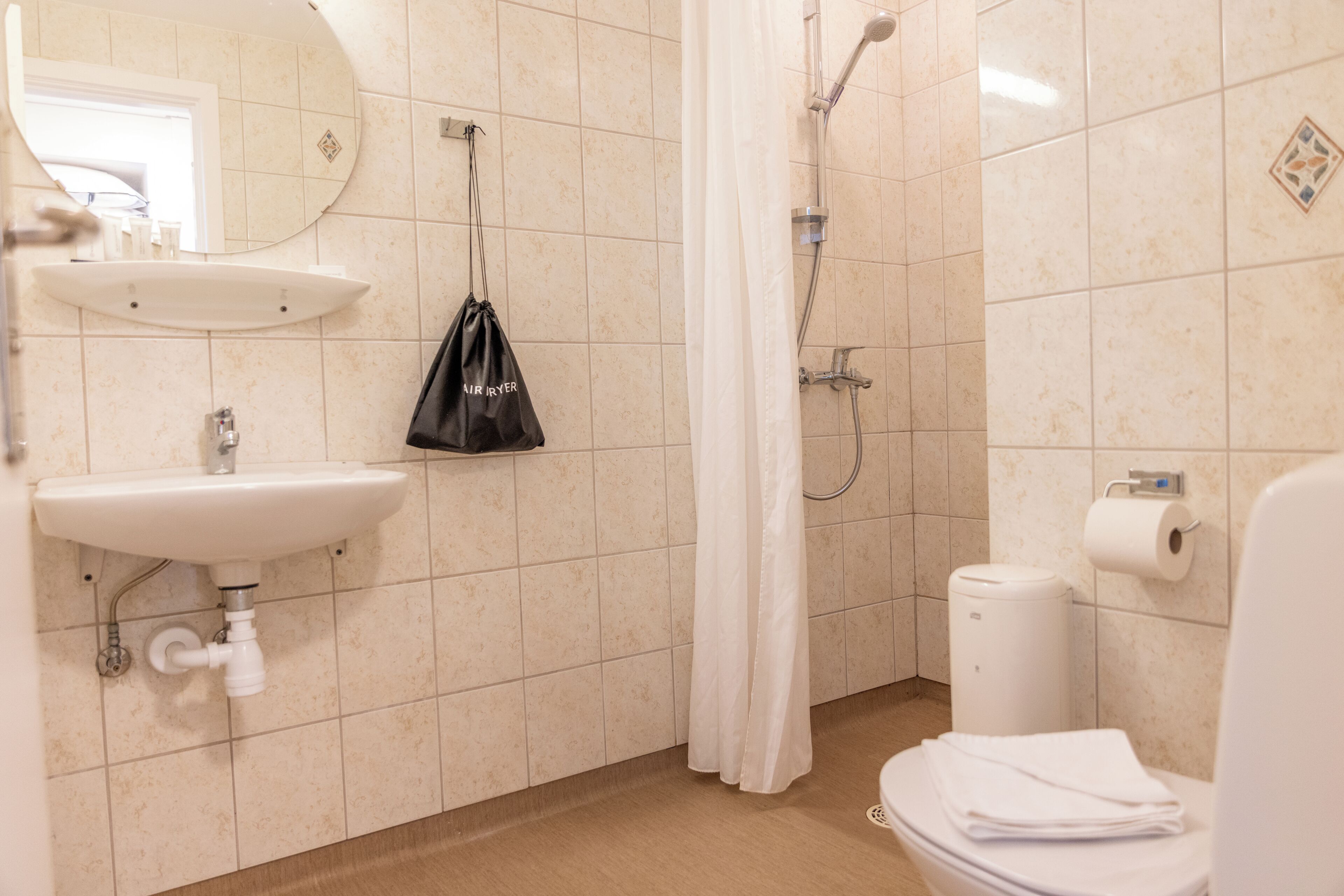 Baño con lavamanos, espejo, ducha, inodoro y una bolsa colgada.