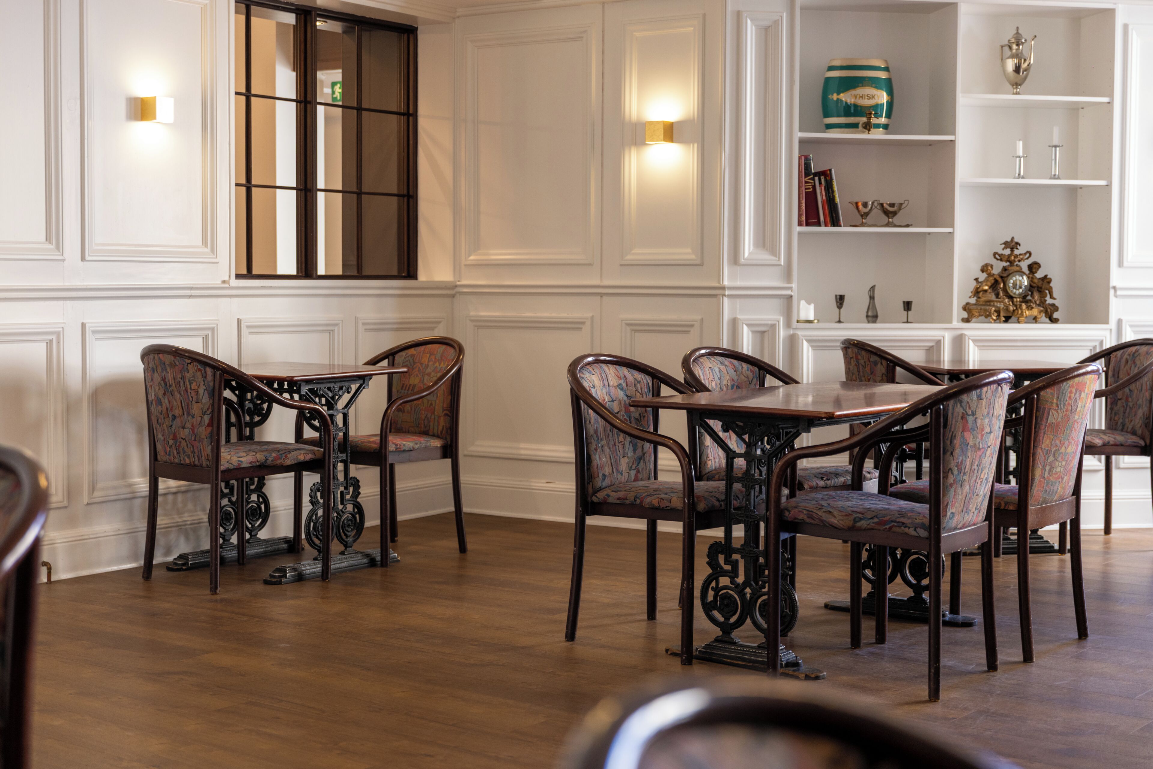 A dining area with a round table and upholstered chairs, a bookshelf with decorative items, and a window with a view.
