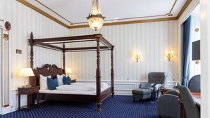 A four-poster bed with a wooden frame, two bedside lamps, a chandelier, a grey armchair, and a small table with a glass top. - Hotel Phønix (Aalborg)