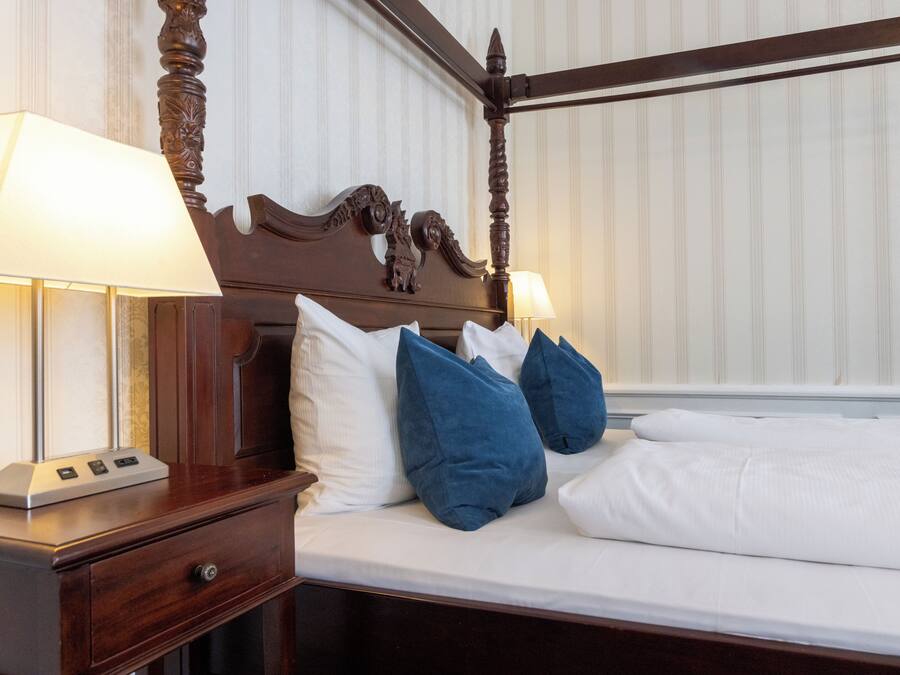 A four-poster bed with white linens, two blue pillows, and a wooden nightstand with a lamp.