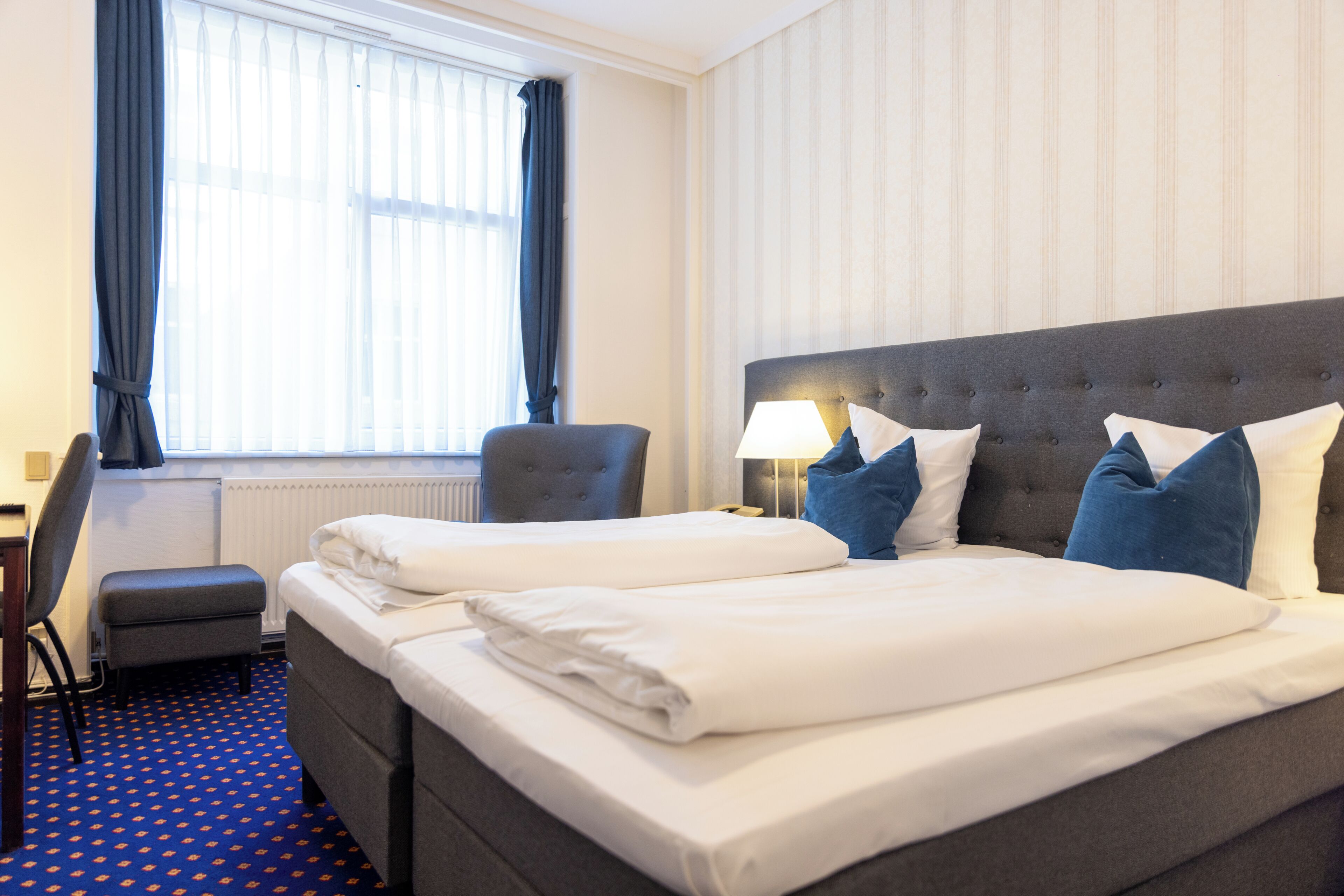 A hotel room with a bed, two chairs, a desk, a lamp, and a window with curtains.