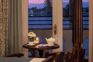 A table with a teapot and cups, a candle, and a vase of flowers, set against a view of a body of water and palm trees. - Brunton Boatyard - Cgh Earth (Kochi)