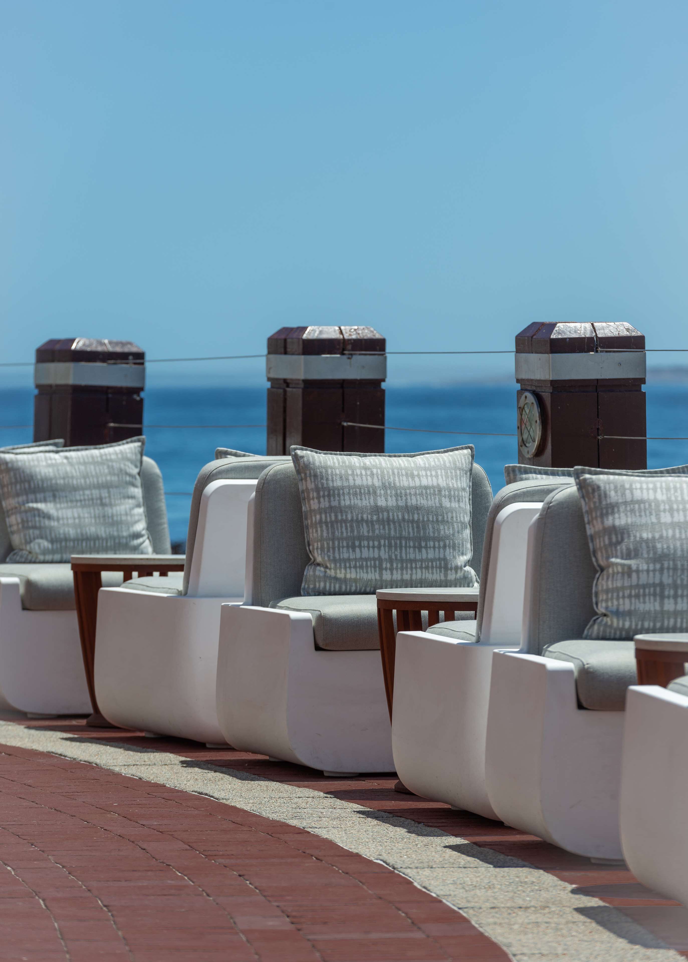 A row of white lounge chairs with grey cushions, positioned on a brick-paved area with a view of the ocean.