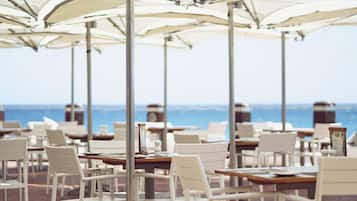 A seaside restaurant with white chairs and tables under large umbrellas, overlooking the ocean.