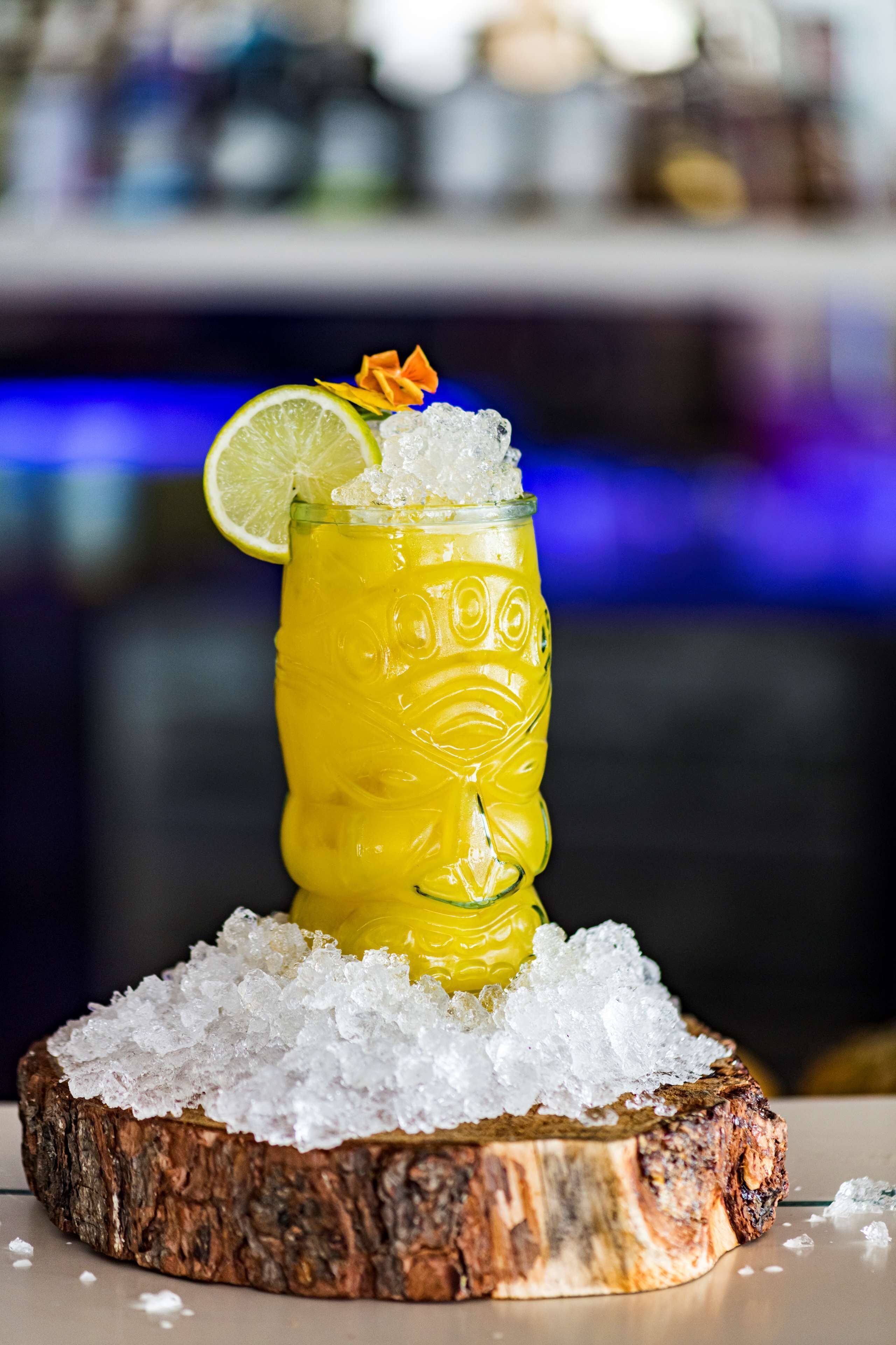 A glass of yellow beverage with ice and a lime slice, placed on a wooden coaster with additional ice.