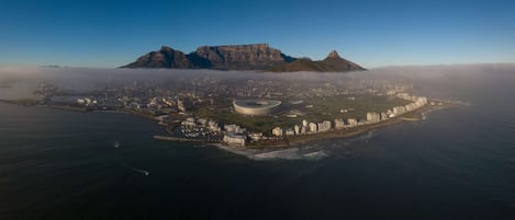 Vista aérea de una ciudad costera con un estadio destacado, edificios residenciales y montañas al fondo.