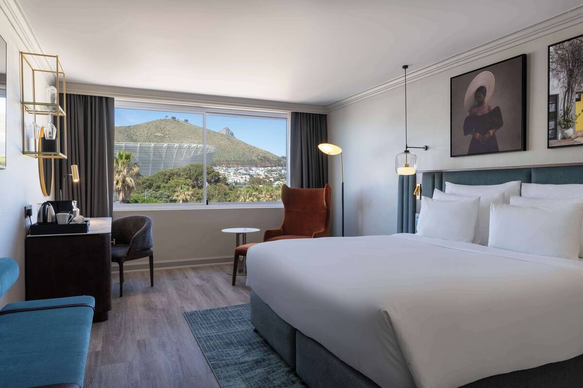 A hotel room with a large bed, a desk with a lamp, a chair, a view of a mountain, and a painting on the wall.