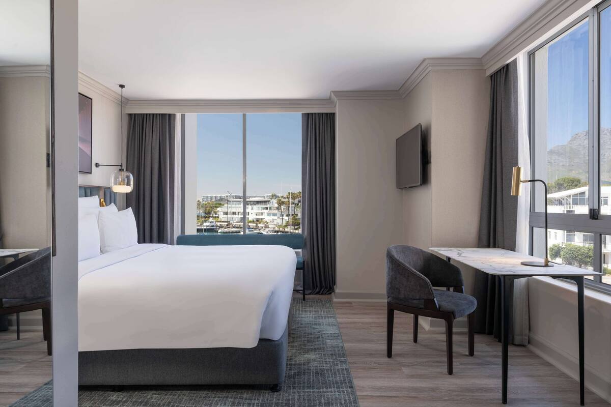 A modern hotel room with a large bed, a desk, and a chair, a TV mounted on the wall, and a view of the city through large windows.