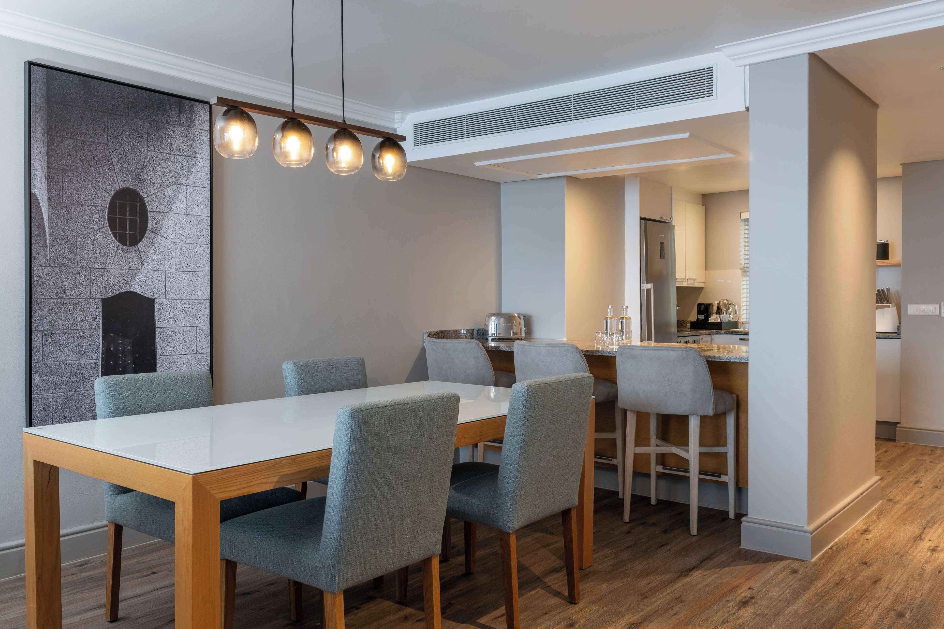 A modern dining area with a white table and grey chairs, a stone wall with a circular window, and a kitchen in the background.