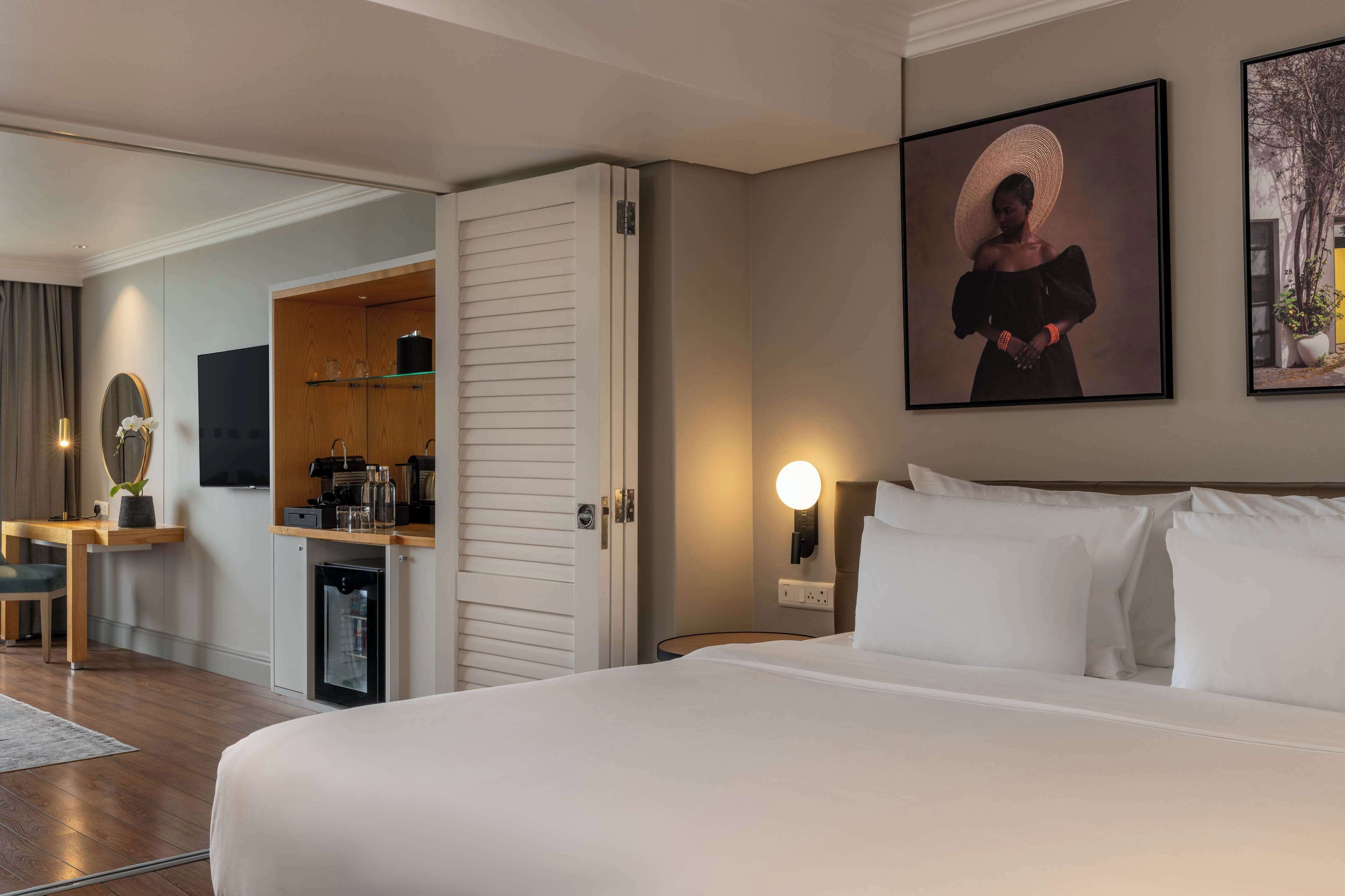 A hotel room with a bed, a desk, a minibar, a television, and artwork on the walls.
