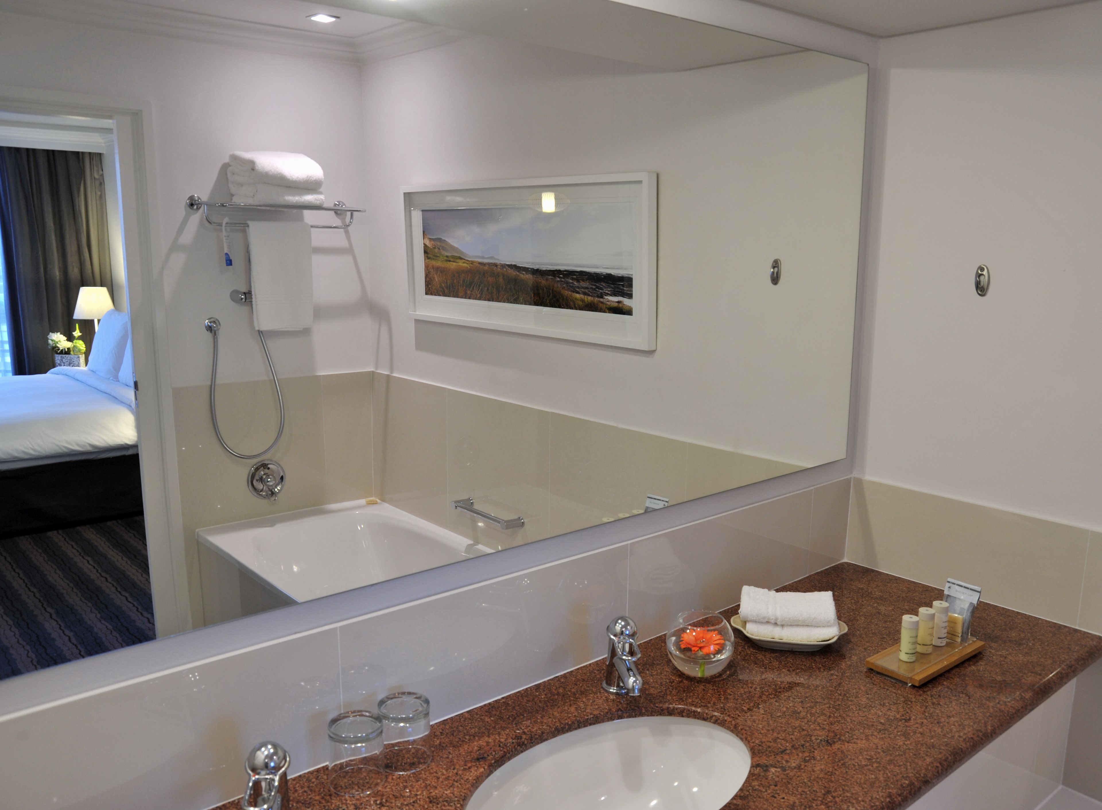 Ein Hotelbadezimmer mit einem großen Spiegel, einem Waschbecken, einer Dusche und einem im Spiegel sichtbaren Bett.