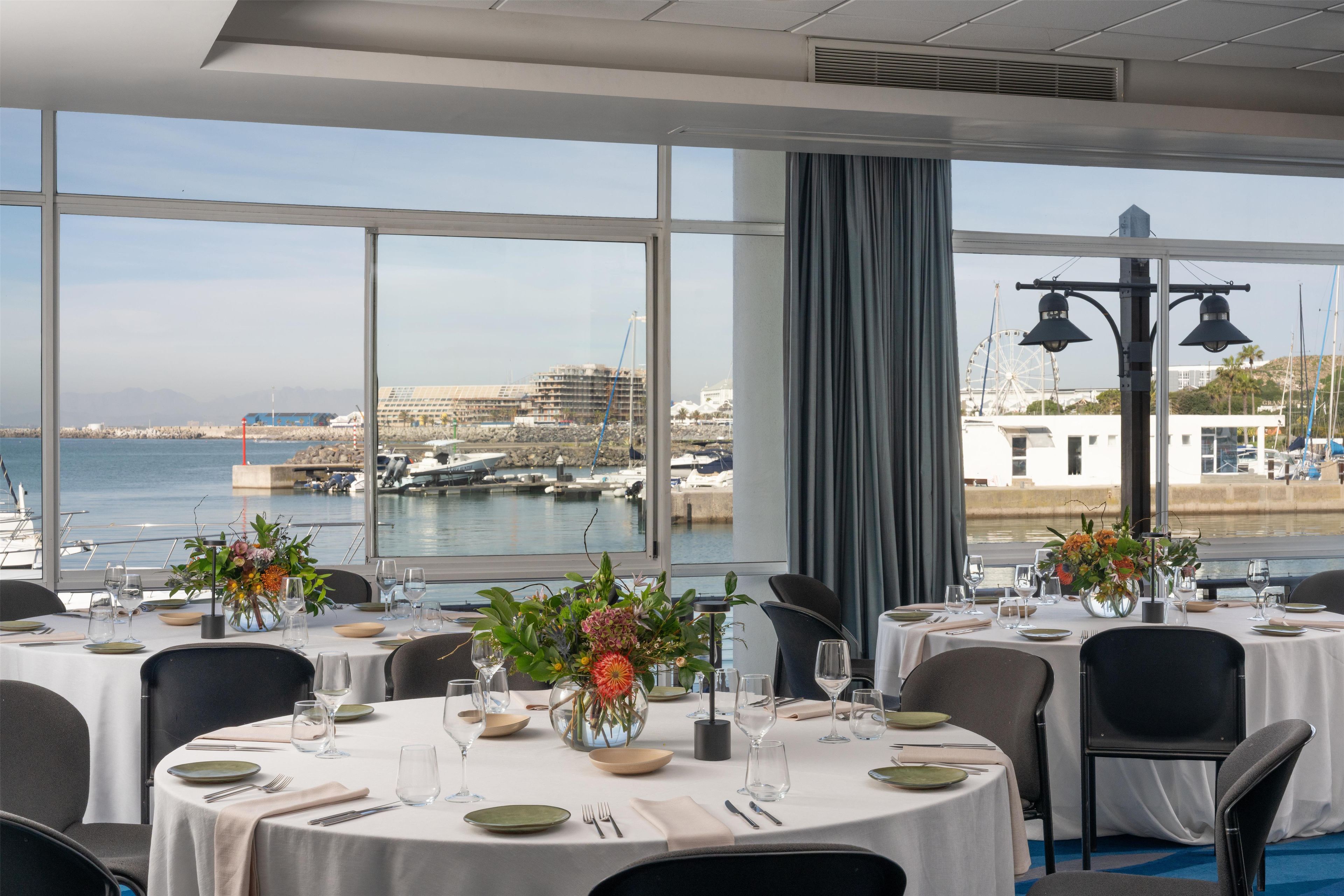 Un espace repas avec des tables dressées pour le service, de grandes fenêtres offrant une vue sur la mer et les bateaux, ainsi qu’une vue sur les bâtiments situés au loin.