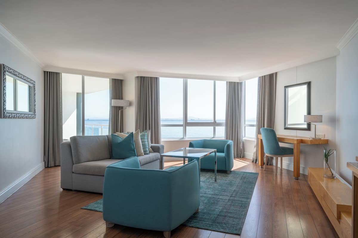 A modern living room with a sofa, two armchairs, a coffee table, and a desk with a chair. Large windows provide a view of the outdoors.