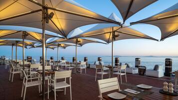 An outdoor dining area with white chairs and tables, large umbrellas, and a view of the sea.