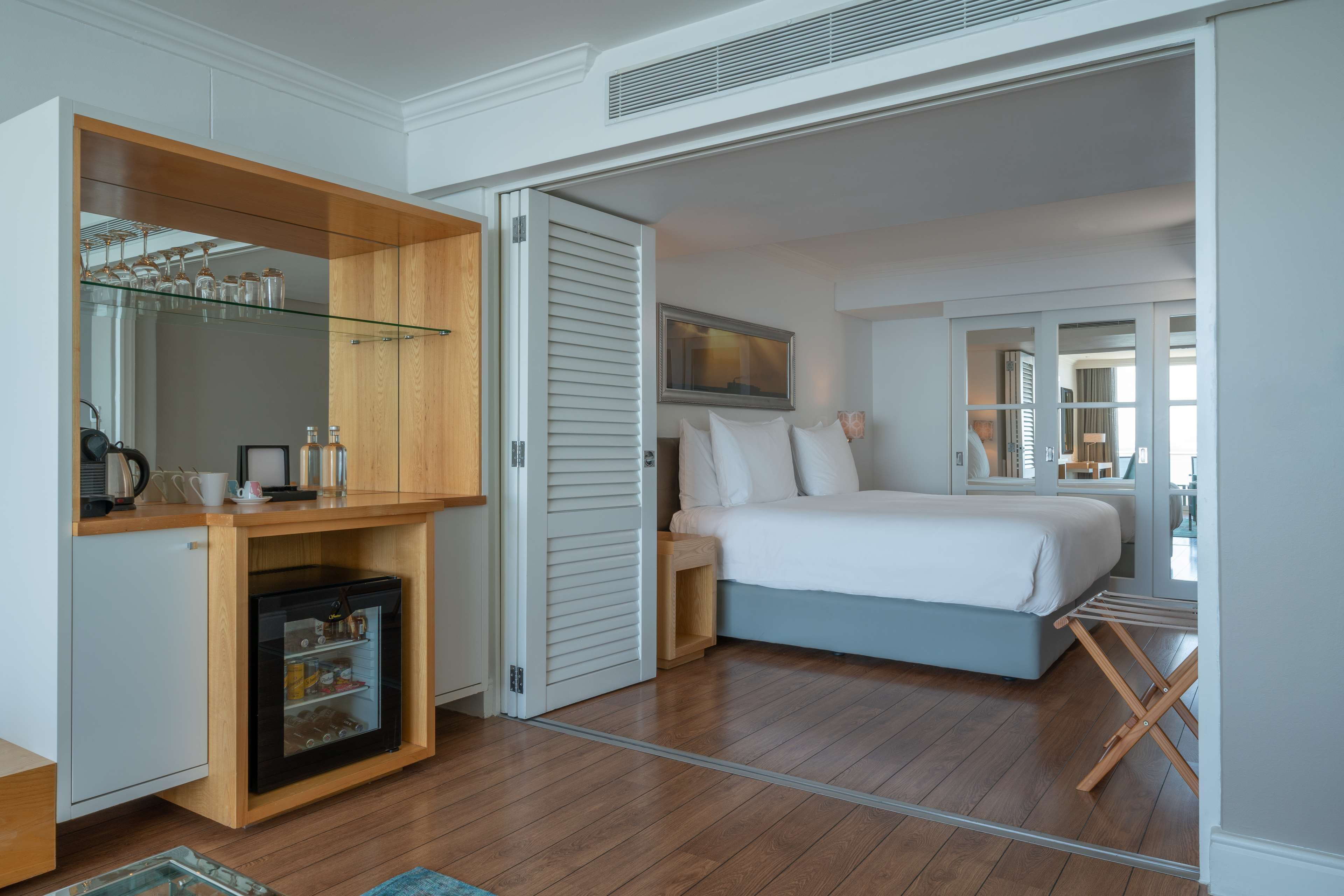 A bedroom with a bed, a small kitchenette, a glass-fronted storage unit, and a sliding door leading to another room.