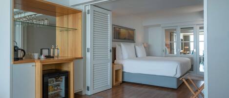 A bedroom with a bed, a small kitchenette, a glass-fronted storage unit, and a sliding door leading to another room.