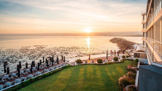 A hotel with a view of the sea at sunset, outdoor seating, and a well-maintained lawn.