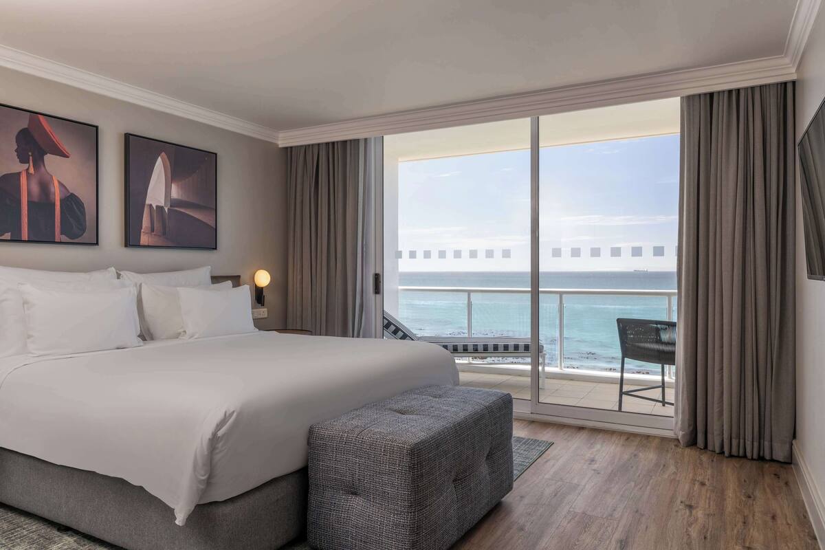 A hotel room with a large bed, a bench, a balcony with a view of the sea, and two framed pictures on the wall.