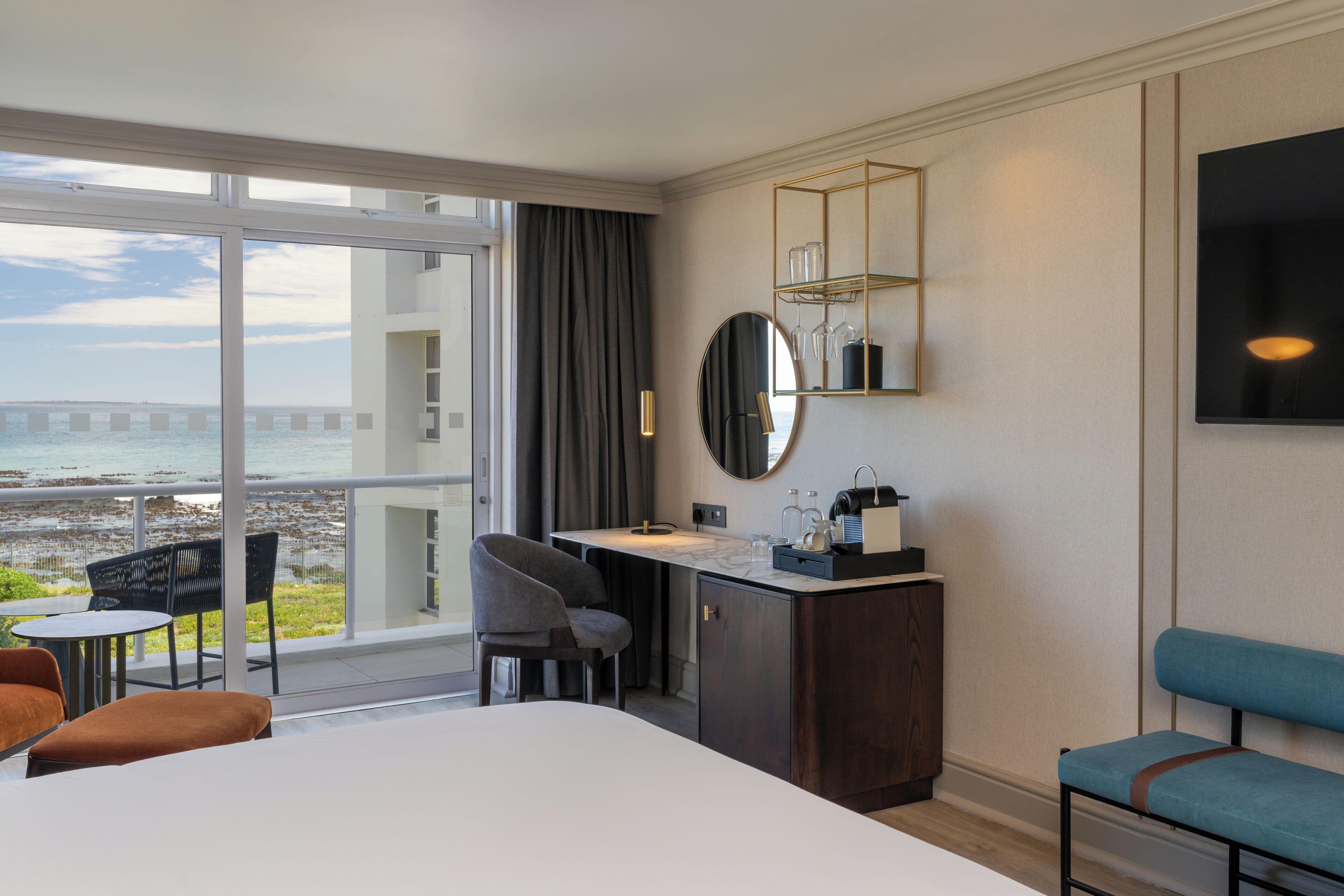 Ett hotellrum med en stor säng, ett skrivbord med en stol, ett litet bord med en stol, en blå bänk, en tv, en spegel, en hylla med glas och utsikt över havet.