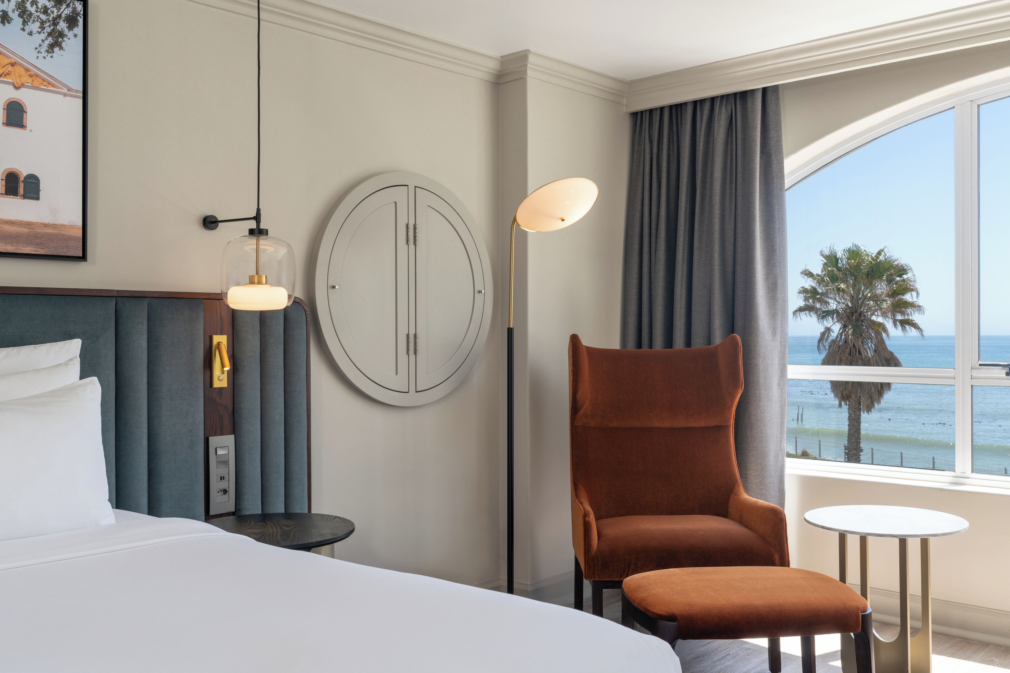 Una habitación de hotel con una cama, una silla, una mesa redonda, una lámpara de pie y vista al mar.