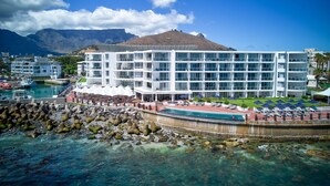 A modern hotel with a pool area, a rocky shoreline, and a mountain in the background.
