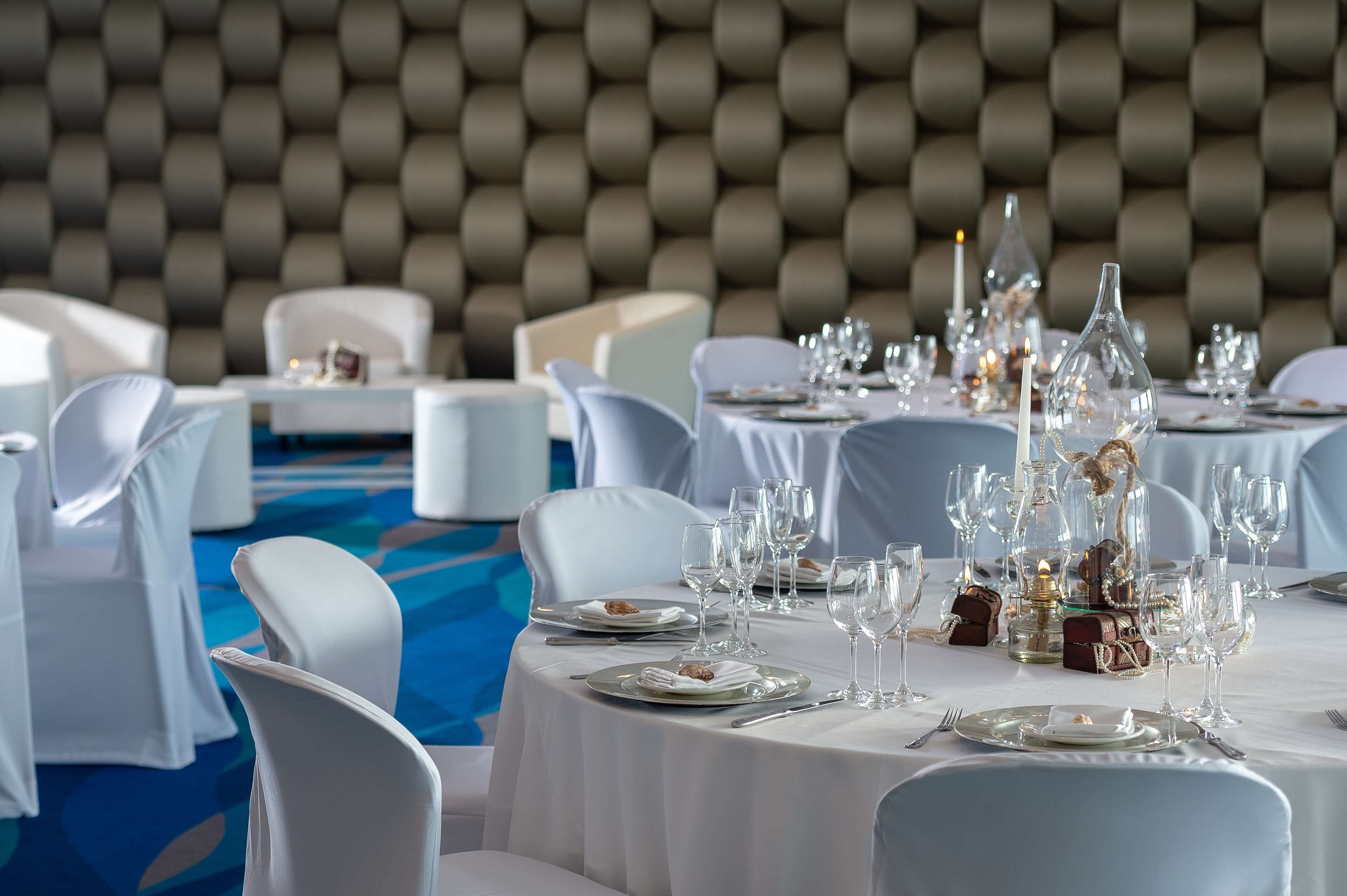 Une salle de banquet avec des tables rondes dressées pour le repas, des chaises blanches et une moquette bleue.