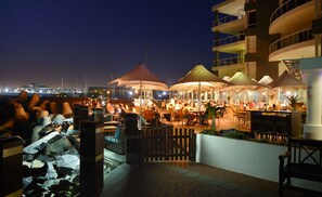 An outdoor dining area with multiple tables and umbrellas, a view of the marina in the background, and a building with balconies. - Radisson Collection Hotel, Waterfront Cape Town (Cape Town)