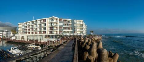 Ein mehrstöckiges Hotel mit Balkonen, eine Marina mit Booten und ein Felsenmole am Meer.